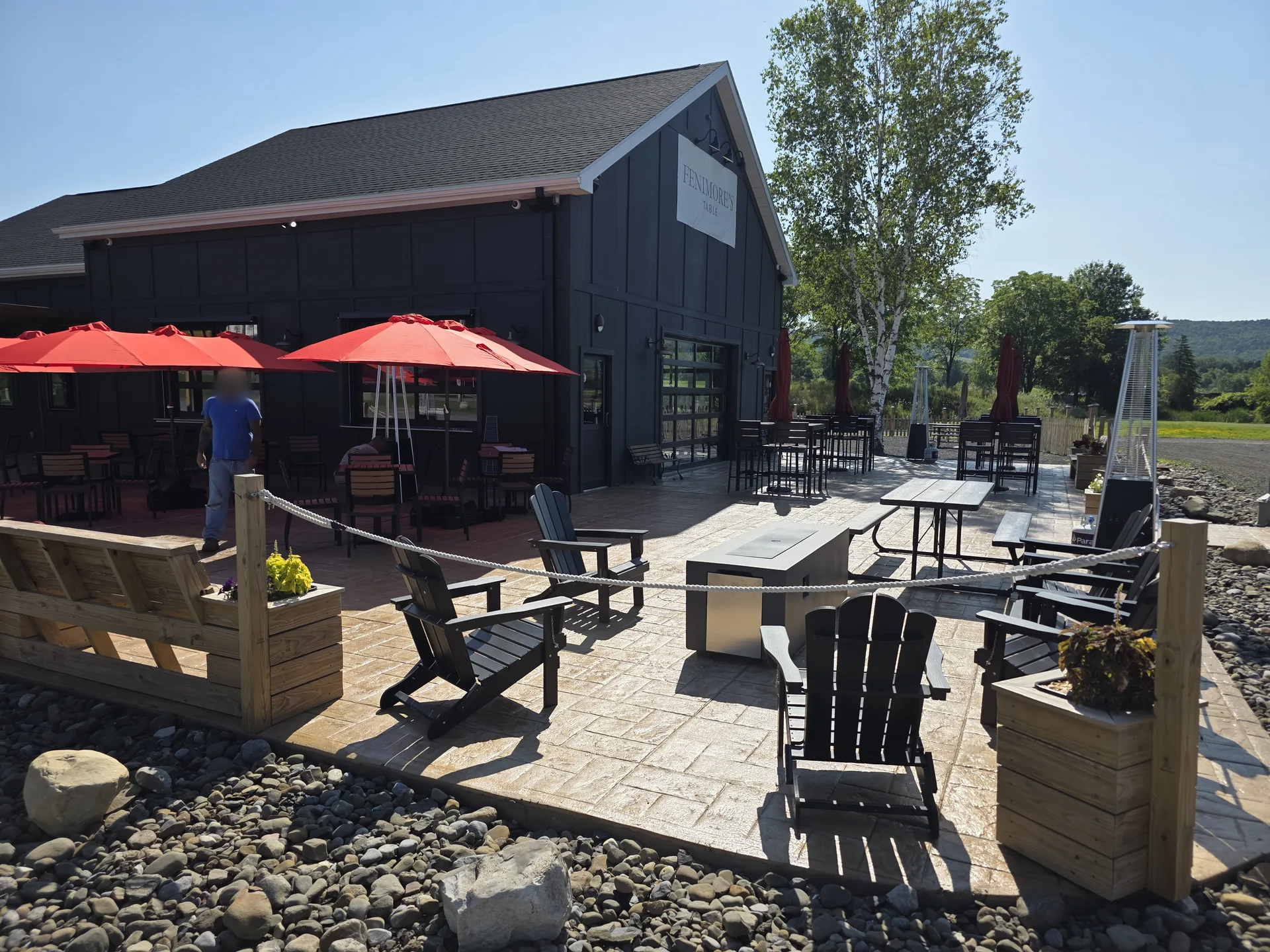 The Fenimore's Table patio on a sunny day, with Adirondack chairs around a fire table, red umbrellas over dining tables and the Fenimore's Table sign on the building The Fenimore's Table patio on a sunny day, with Adirondack chairs around a fire table, red umbrellas over dining tables and the Fenimore's Table sign on the building