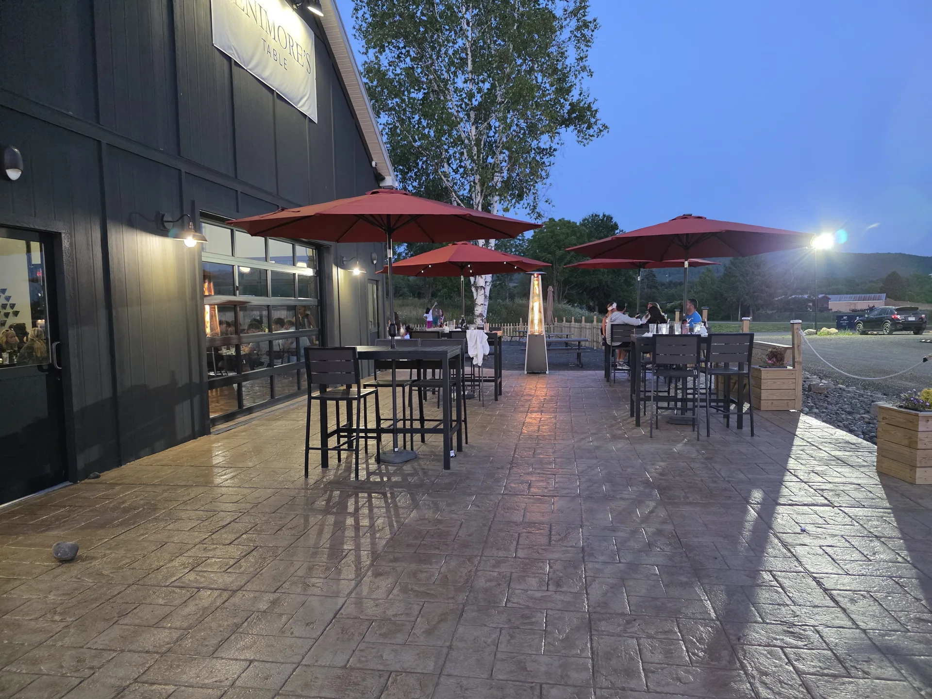 Guests at high-top tables on the Fenimore's Table patio at dusk, under red umbrellas beside the building's glass garage doors and Fenimore's Table sign Guests at high-top tables on the Fenimore's Table patio at dusk, under red umbrellas beside the building's glass garage doors and Fenimore's Table sign