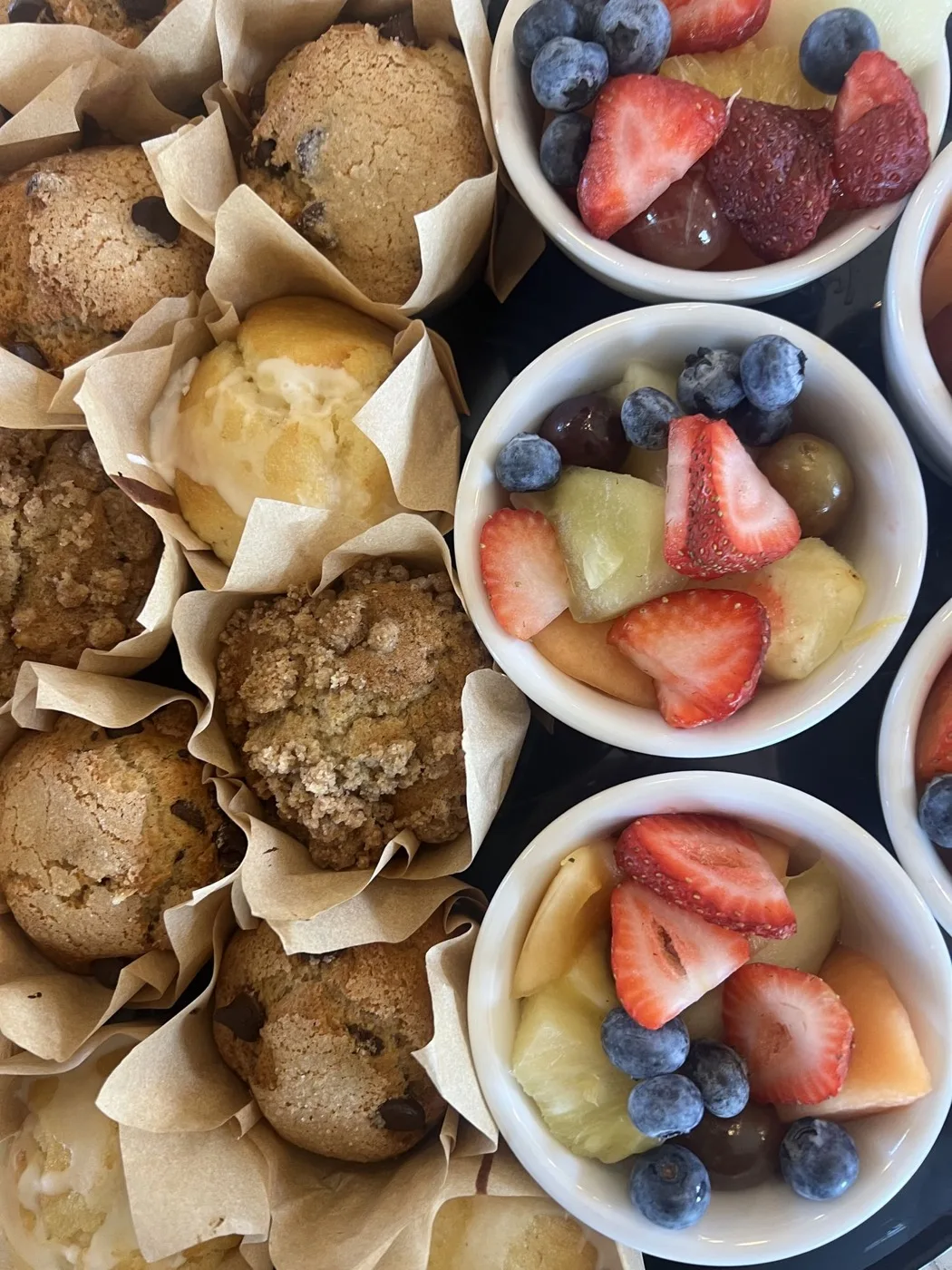 Muffins in paper liners next to bowls of strawberries, blueberries, grapes, melon and pineapple Muffins in paper liners next to bowls of strawberries, blueberries, grapes, melon and pineapple