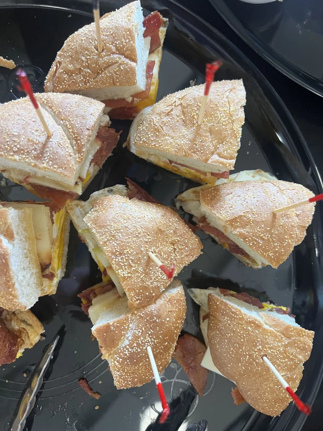 A black platter of halved sandwiches on seeded rolls, held with red cocktail picks A black platter of halved sandwiches on seeded rolls, held with red cocktail picks