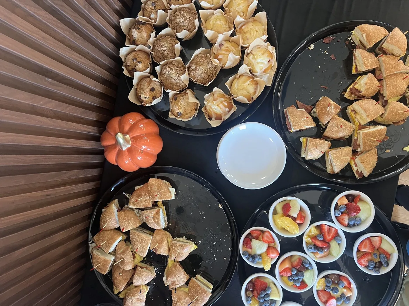 A Fenimore's Table catering spread on a dark table: platters of muffins and cut sandwiches, fruit cups and a small orange pumpkin A Fenimore's Table catering spread on a dark table: platters of muffins and cut sandwiches, fruit cups and a small orange pumpkin